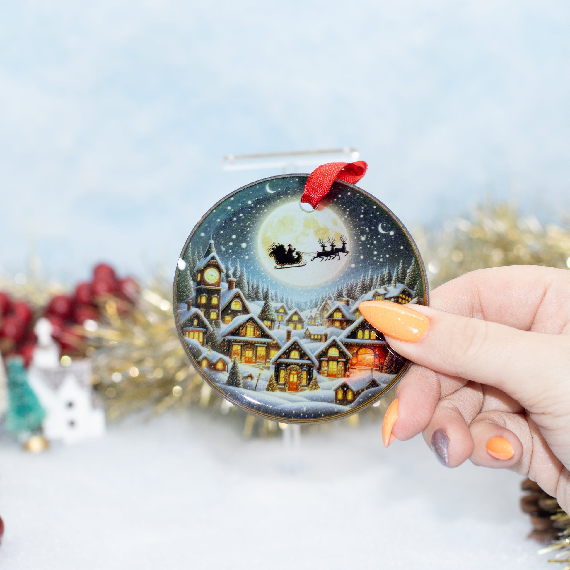 A hand holds a round Christmas ornament featuring a painted snowy village and Santa Claus in the sky. The ornament has a red hanging ribbon, and the background is softly blurred with festive decorations.