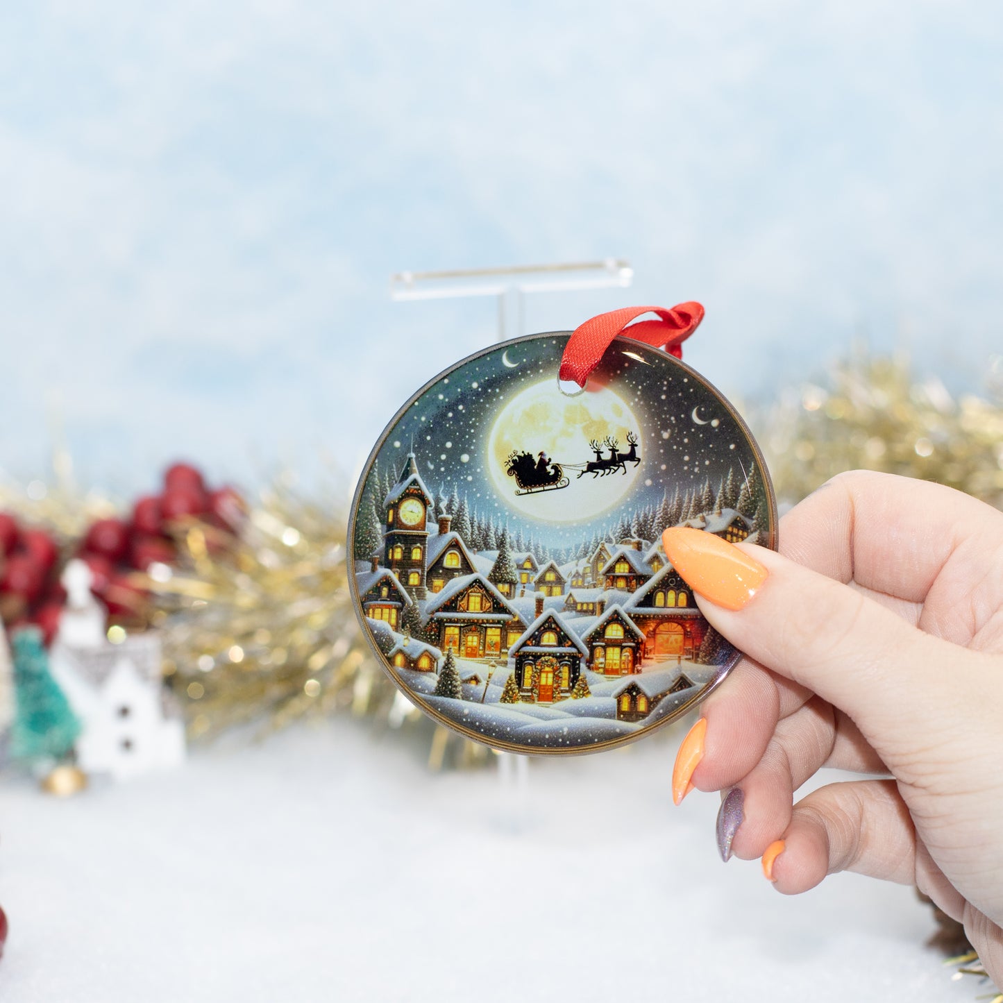 A hand holds a round Christmas ornament featuring a snowy village, glowing houses, Santa Claus in his sleigh, and a red hanging ribbon. The background is a soft, out-of-focus holiday scene with gold tinsel and red berries.