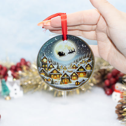 A hand holds a round Christmas ornament featuring a snowy village at night with Santa Claus flying in his sleigh under a full moon. The ornament has a red hanging ribbon and is photographed against a softly blurred festive background with tinsel and berries.