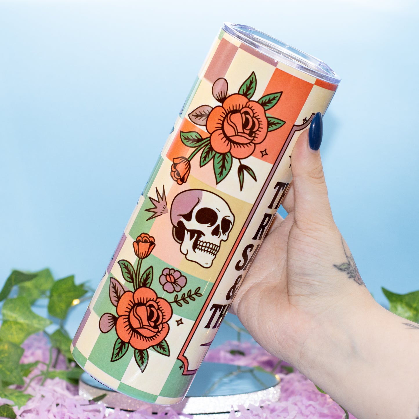 A hand holds a tall insulated tumbler featuring a retro tattoo-style design with orange roses, a skull, and checkered blocks on a cream background. The tumbler has a clear lid and is photographed against a light blue backdrop with decorative green leaves and pink petals at the bottom.