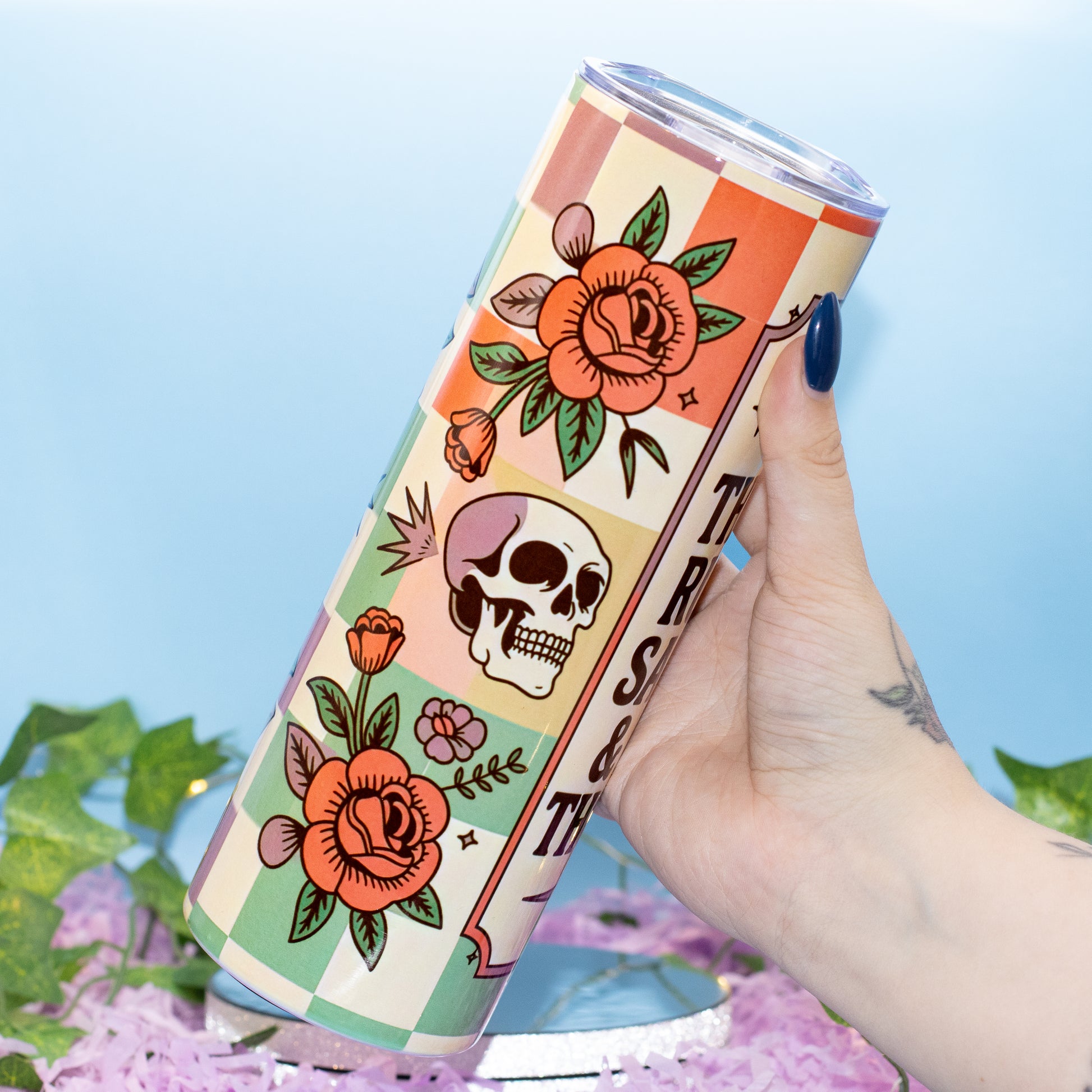 A hand holds a tall insulated tumbler featuring a retro tattoo-style design with orange roses, a skull, and checkered blocks on a cream background. The tumbler has a clear lid and is photographed against a light blue backdrop with decorative green leaves and pink petals at the bottom.