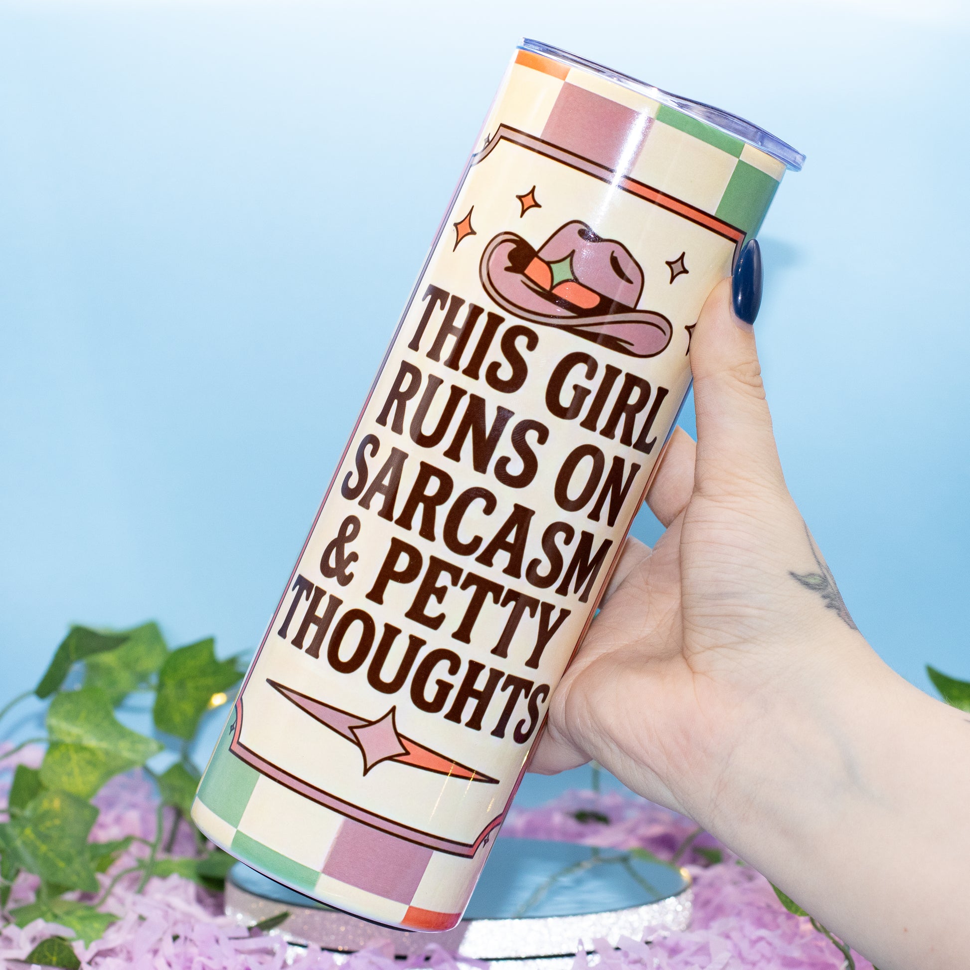 A hand holds a tall insulated tumbler with a printed western-themed design. The tumbler has a cream background with brown text reading 'THIS GIRL RUNS ON SARCASM & PETTY THOUGHTS,' a pink cowboy hat illustration, and pastel checkered bands at the top and bottom.