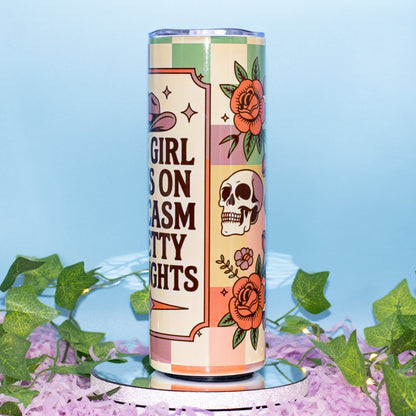 A tall insulated tumbler with a retro checkerboard design in pastel pink, green, and cream, featuring illustrations of a skull, roses, and a cowboy hat. The tumbler is displayed on a mirrored pedestal surrounded by purple flower petals and green ivy against a light blue background.