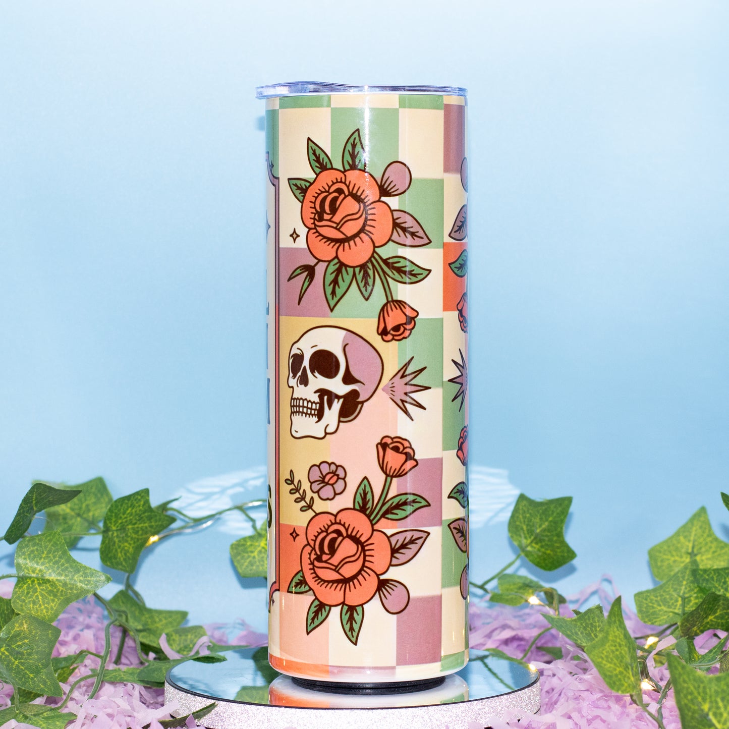 A tall insulated tumbler with a retro checkerboard pattern and floral/illustrated motifs, displayed on a mirrored base with artificial ivy and purple petals against a light blue background.