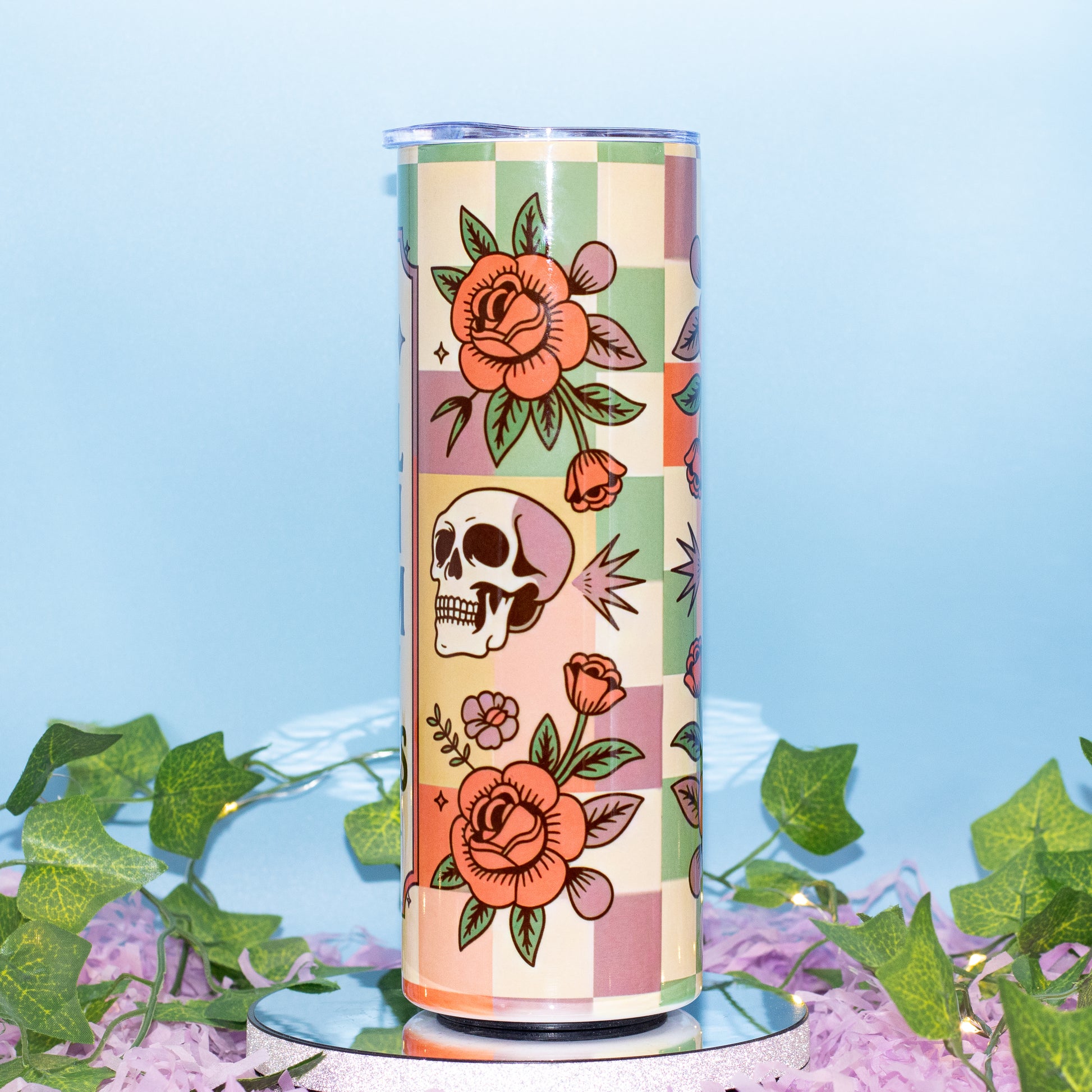 A tall insulated tumbler with a retro checkerboard pattern and floral/illustrated motifs, displayed on a mirrored base with artificial ivy and purple petals against a light blue background.