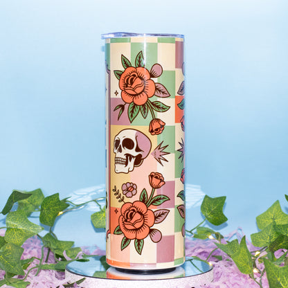 A tall insulated tumbler with a retro checkerboard pattern and floral/illustrated motifs, displayed on a mirrored base with artificial ivy and purple petals against a light blue background.