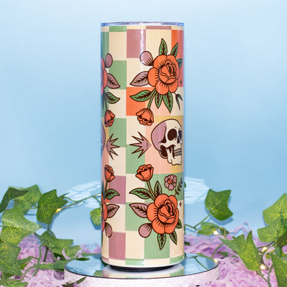 A tall cylindrical tumbler with a retro checkerboard pattern in pastel pink, green, and beige, decorated with orange roses, green leaves, and a skull. The tumbler is displayed on a mirrored surface with artificial green ivy and purple flower petals against a light blue background.