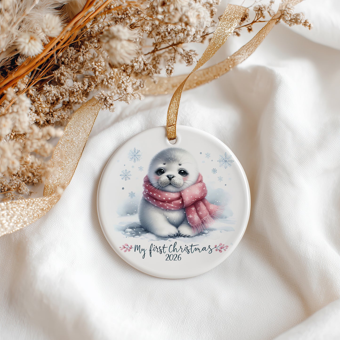 A round ceramic Christmas ornament featuring a cute illustration of a baby seal wearing a pink scarf in the snow, with the text 'My first Christmas 2026' printed below. The ornament hangs from a gold ribbon and is styled on white fabric with dried flowers and a gold ribbon.