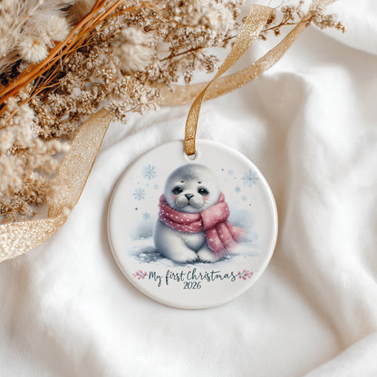 A round ceramic Christmas ornament featuring a cute illustration of a baby seal wearing a pink scarf in the snow, with the text 'My first Christmas 2026' printed below. The ornament hangs from a gold ribbon and is styled on white fabric with dried flowers and a gold ribbon.