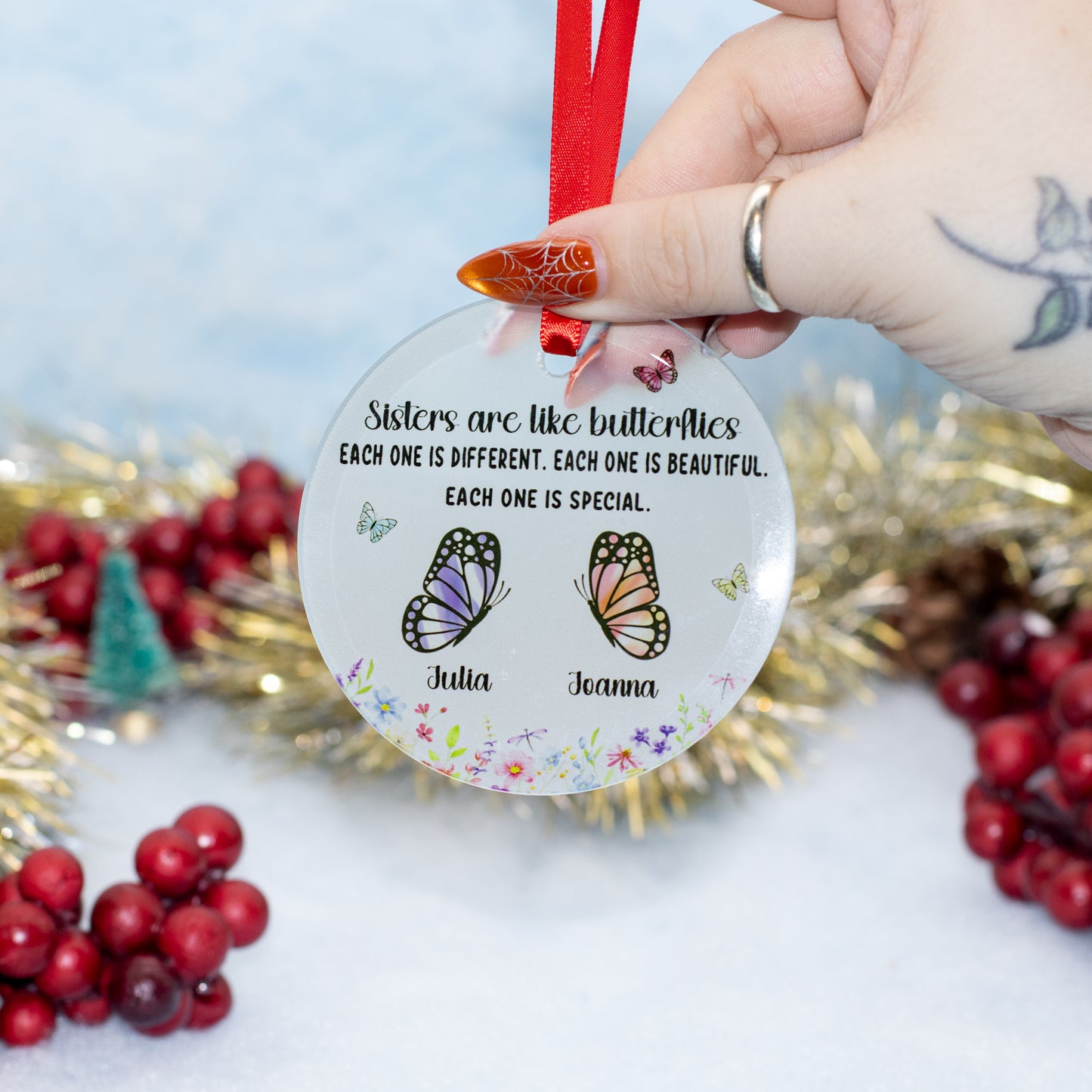 A hand holds a round clear personalized Christmas ornament with a red ribbon. The ornament features black text reading 'Sisters are like butterflys EACH ONE IS DIFFERENT.