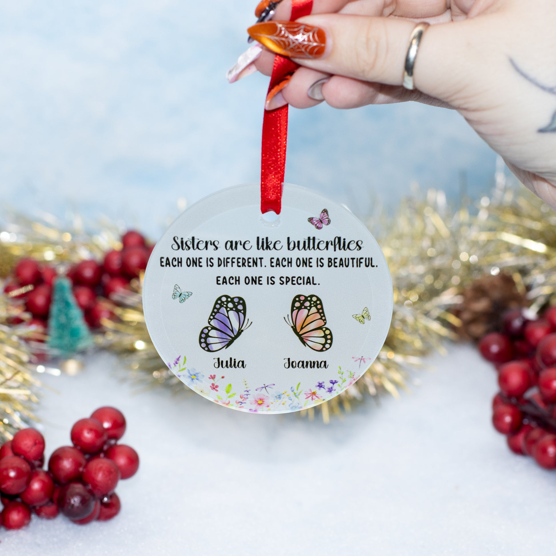 A hand holds a round white personalised ornament with a red ribbon. The ornament features a printed quote about butterflies and two butterfly illustrations with the names Julia and Jenna.