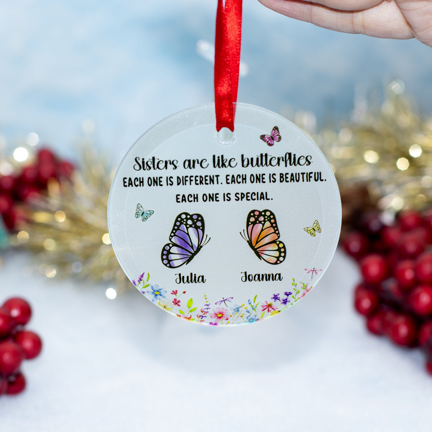 A hand holds a round clear personalized Christmas ornament with a red ribbon. The ornament features black text reading 'Sisters are like butterflies EACH ONE IS DIFFERENT.