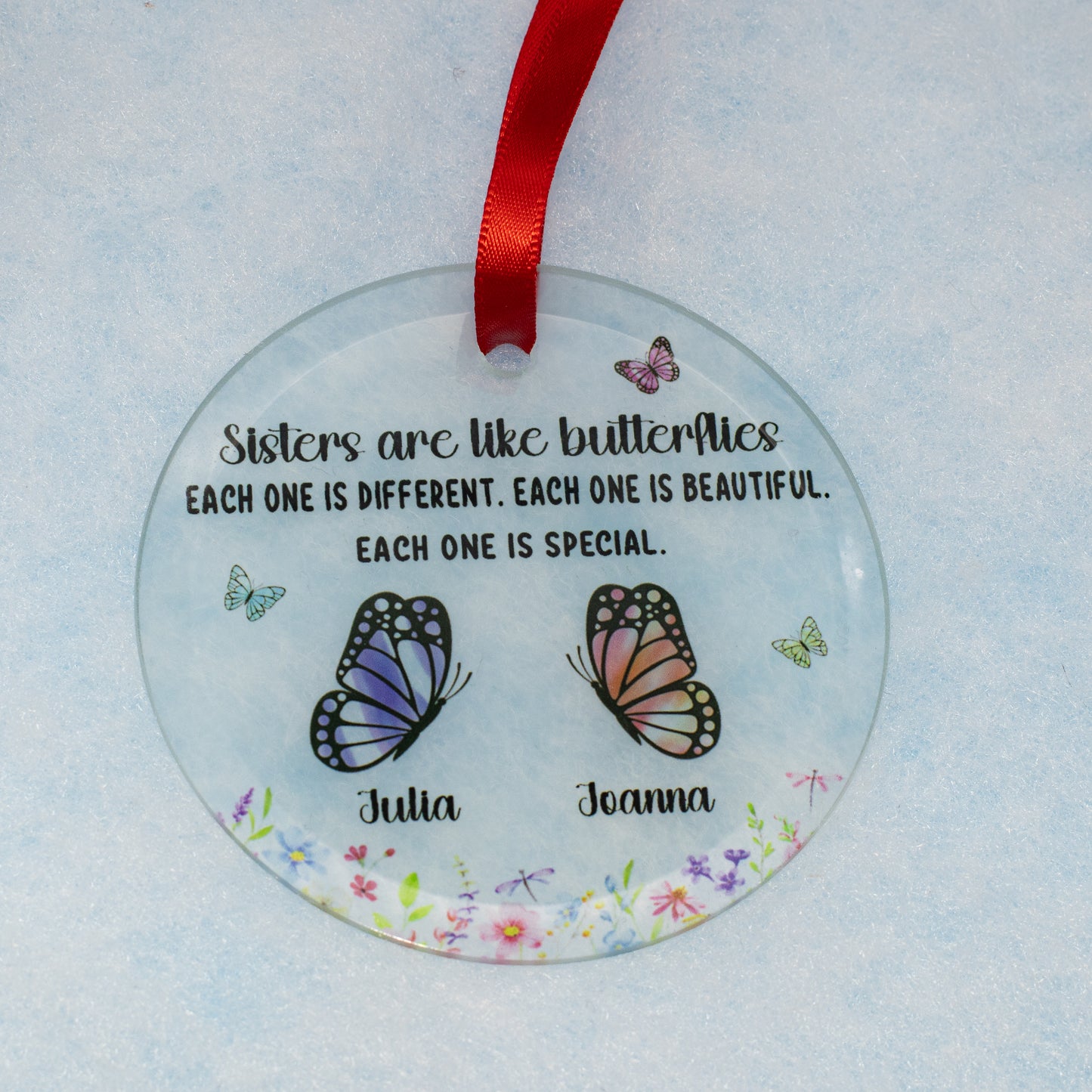A personalized round clear glass ornament featuring a red ribbon at the top, printed with the quote 'Sisters are like butterflies' and two illustrated butterflies labeled 'Julia' and 'Joanna' on a light blue textured background.
