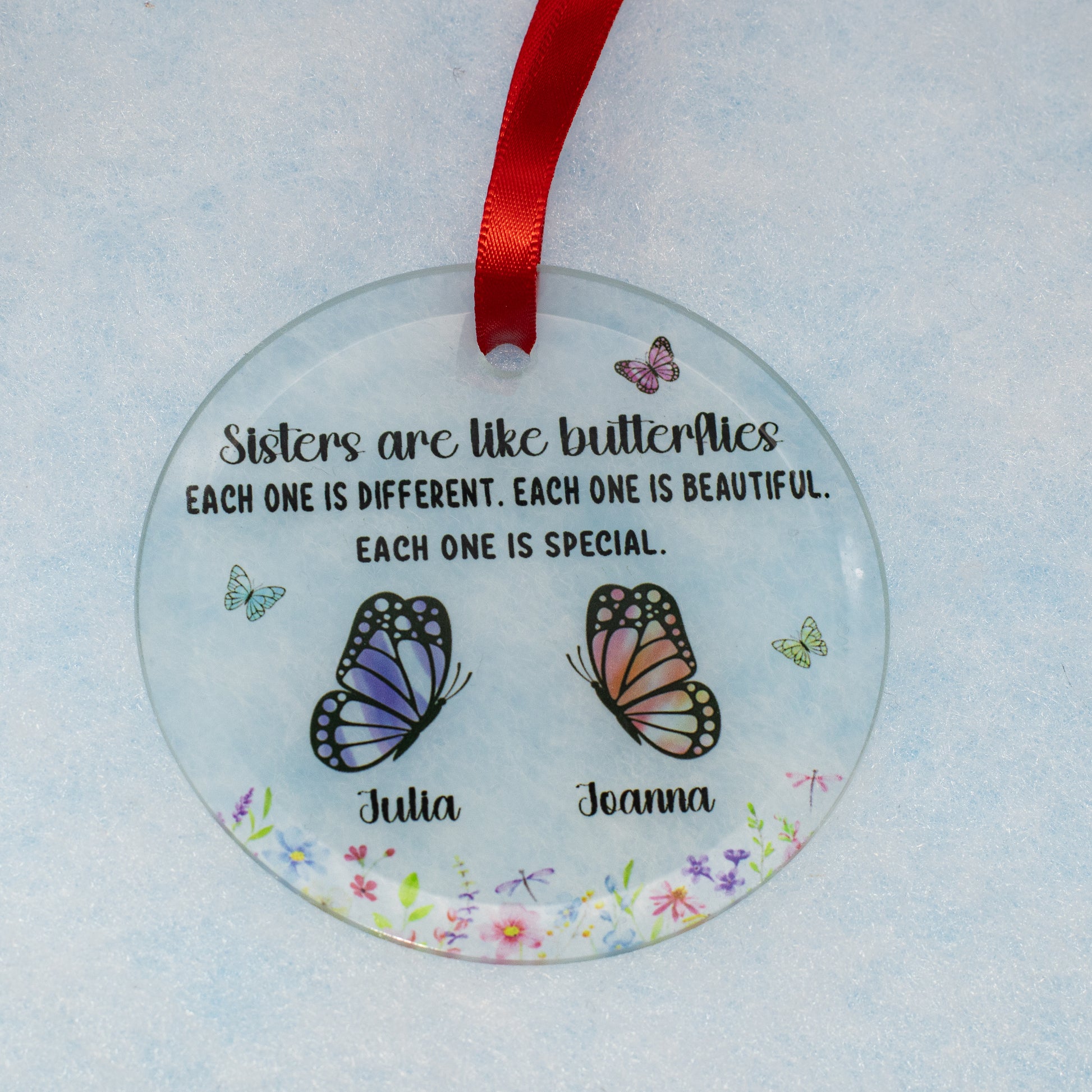 A personalized round clear glass ornament featuring a red ribbon at the top, printed with the quote 'Sisters are like butterflies' and two illustrated butterflies labeled 'Julia' and 'Joanna' on a light blue textured background.