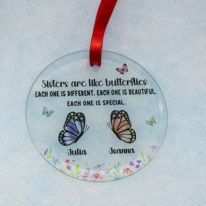 A personalized round clear glass ornament featuring a red ribbon at the top, printed with the quote 'Sisters are like butterflies' and two illustrated butterflies labeled 'Julia' and 'Joanna' on a light blue textured background.