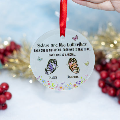 A personalised round clear glass Christmas ornament hanging from a red ribbon, held by a hand. The ornament features a printed message about sisters being like butterflies, two illustrated butterflies (one purple, one orange), and the names 'Julia' and 'Joanna' at the bottom.