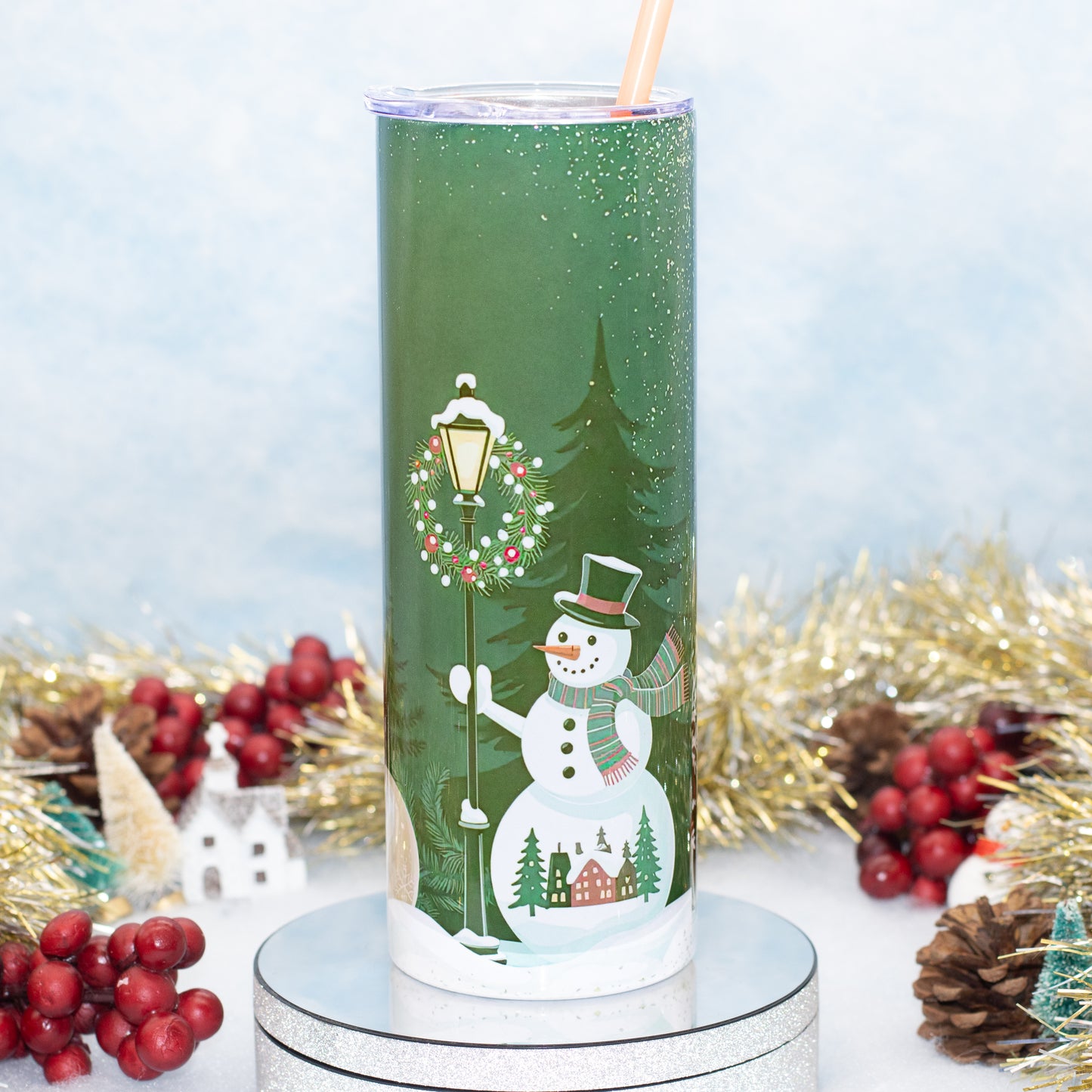 A tall insulated tumbler with a clear lid and straw, decorated with a festive winter scene featuring a snowman, evergreen trees, a street lamp, and small houses. The tumbler is displayed on a round silver pedestal with holiday decorations like red berries, pinecones, and gold tinsel in the background.