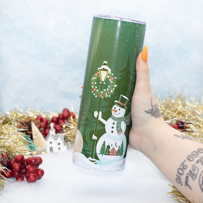 A hand holds a green insulated tumbler decorated with a winter scene featuring a snowman, Christmas wreath, street lamp, evergreen trees, and small houses. The background is decorated with gold tinsel, red berries, and a soft light blue backdrop.