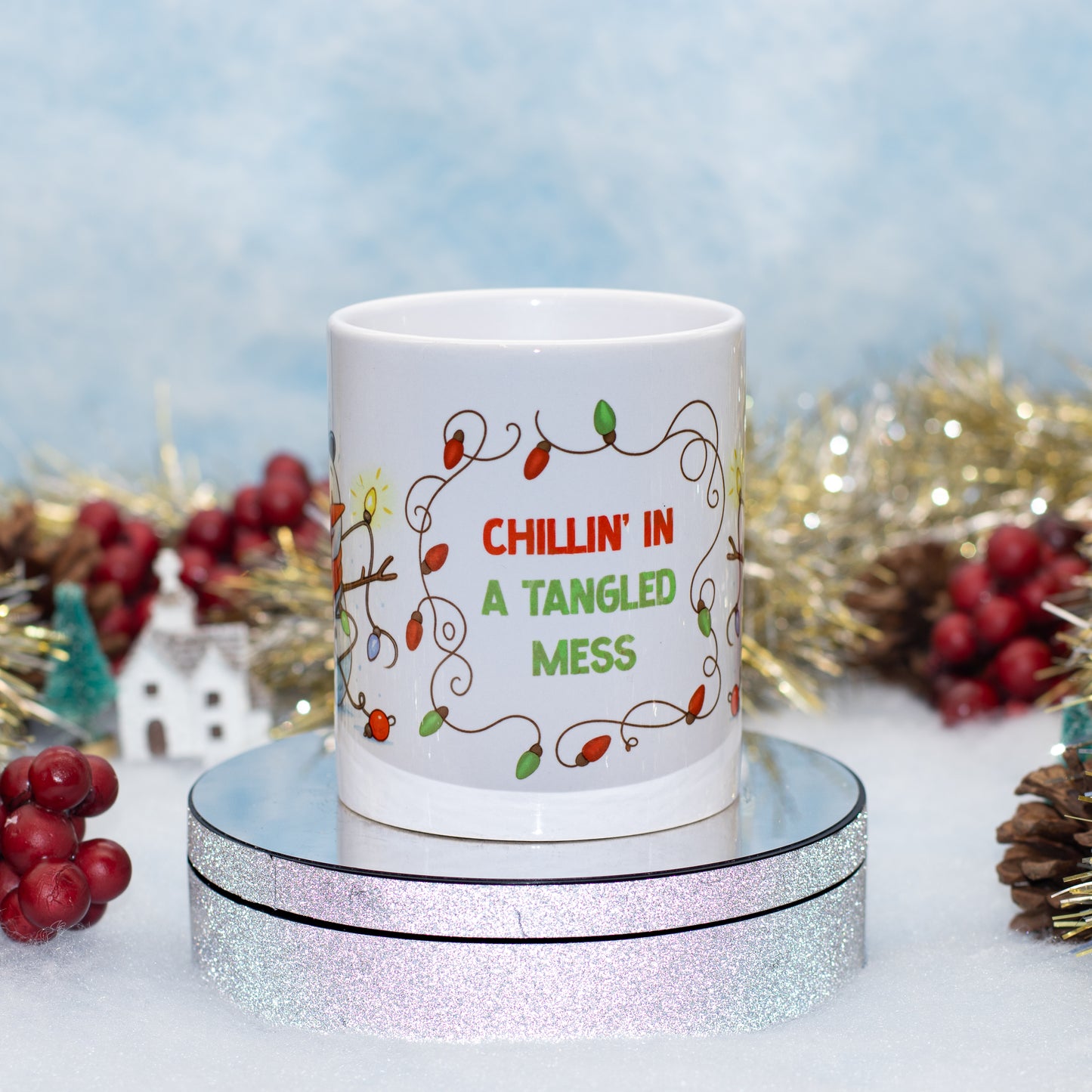 A white ceramic mug with a Christmas holiday design, sitting on a round silver glitter display stand. The mug features a string of colorful lights and the text 'CHILLIN' IN A TANGLED MESS' in red and green.