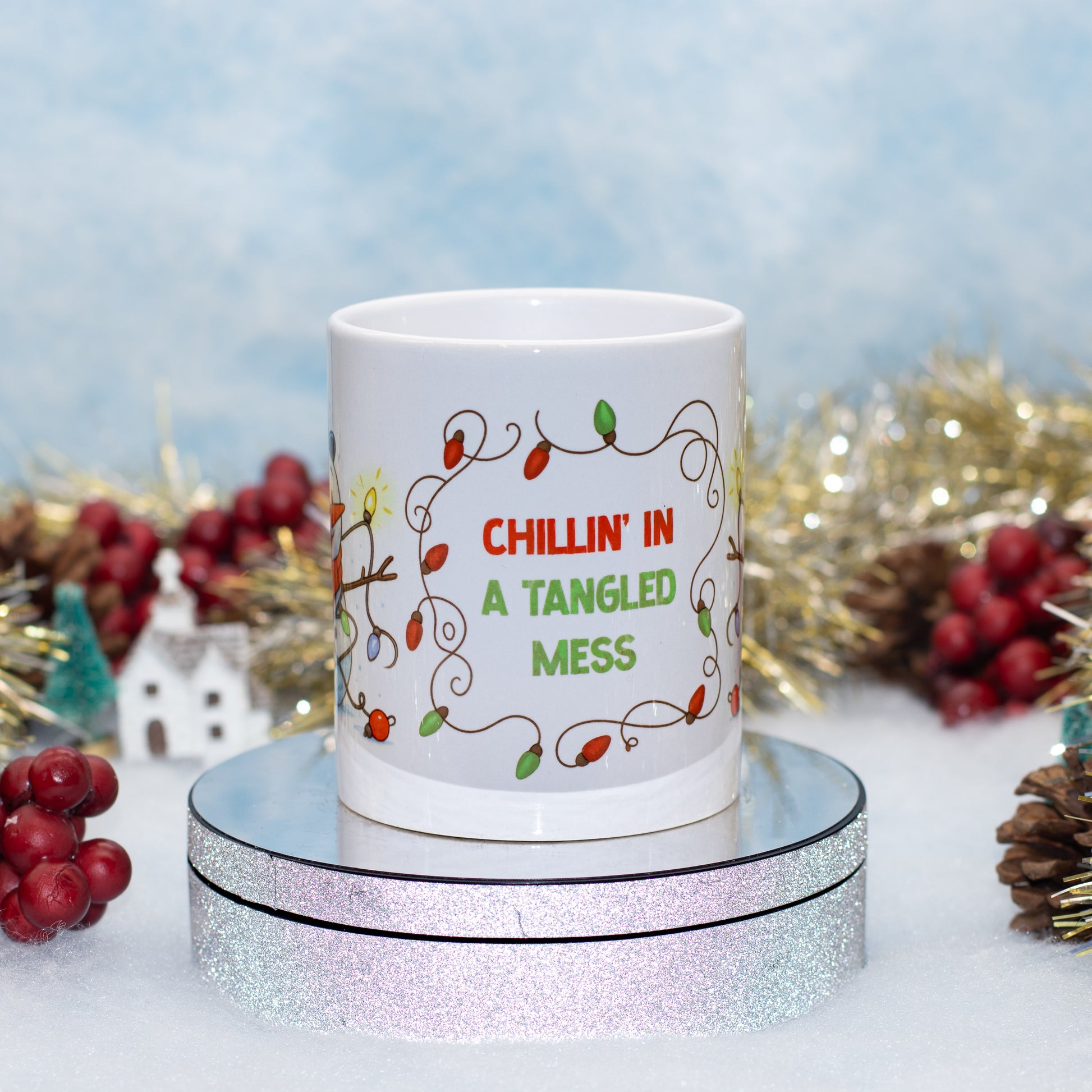 A white ceramic mug with a Christmas holiday design, sitting on a round silver glitter display stand. The mug features a string of colorful lights and the text 'CHILLIN' IN A TANGLED MESS' in red and green.