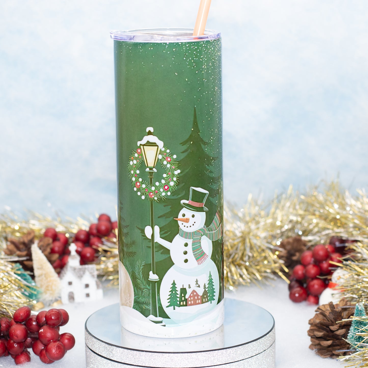 A tall green tumbler with a clear lid and straw, decorated with a winter holiday scene featuring a snowman, street lamp, evergreen tree, and small houses. The tumbler is displayed on a silver pedestal with festive holiday decorations in the background.