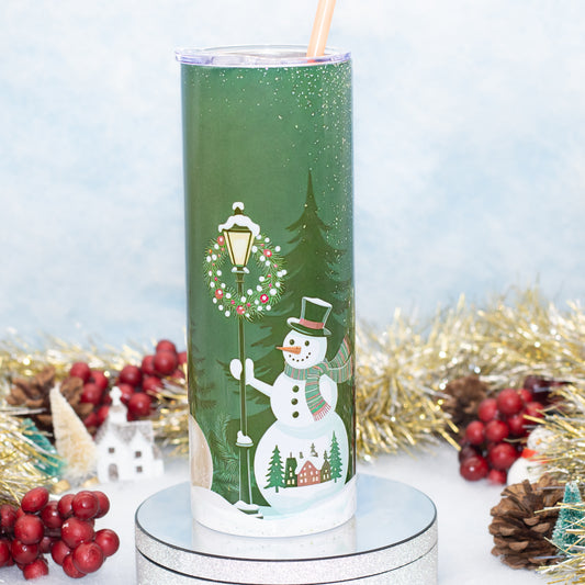 A tall green tumbler with a clear lid and straw, decorated with a winter holiday scene featuring a snowman, street lamp, evergreen tree, and small houses. The tumbler is displayed on a silver pedestal with festive holiday decorations in the background.