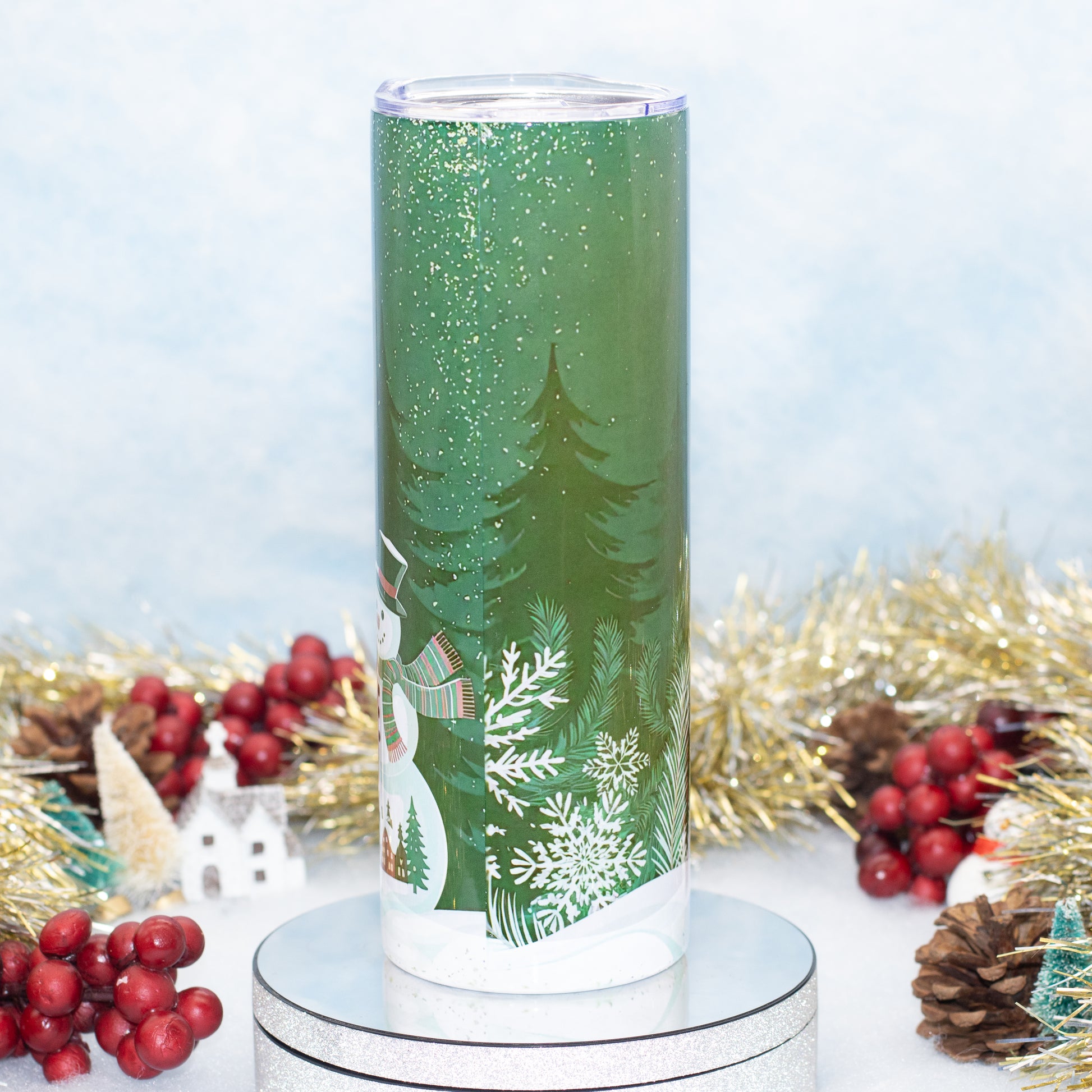 A tall green insulated tumbler with a festive winter forest design, featuring pine trees, snowflakes, and a snowman, shown on a silver pedestal with holiday decorations in the background.