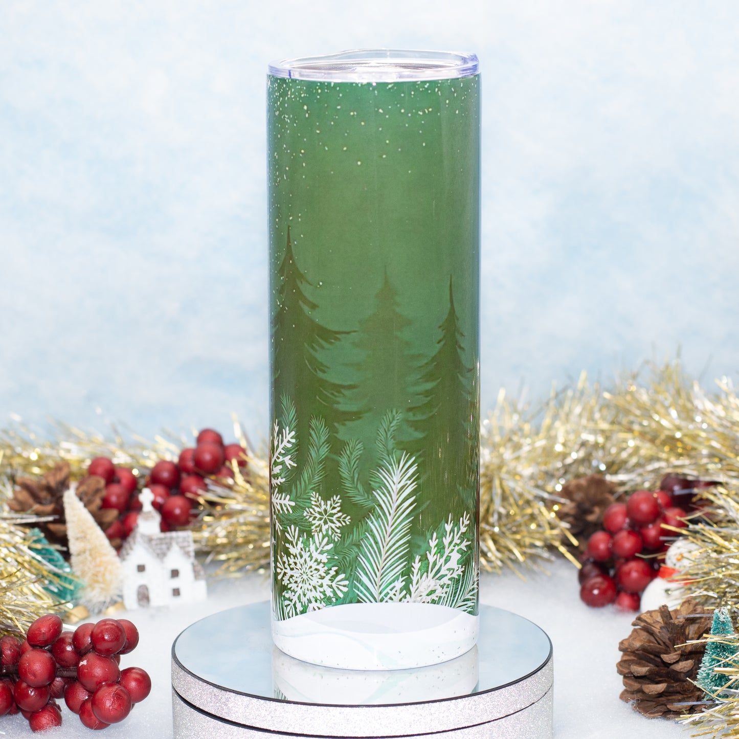 A tall green insulated tumbler with a winter forest design, featuring white pine trees and snowflakes, displayed on a silver pedestal with holiday decorations in the background.