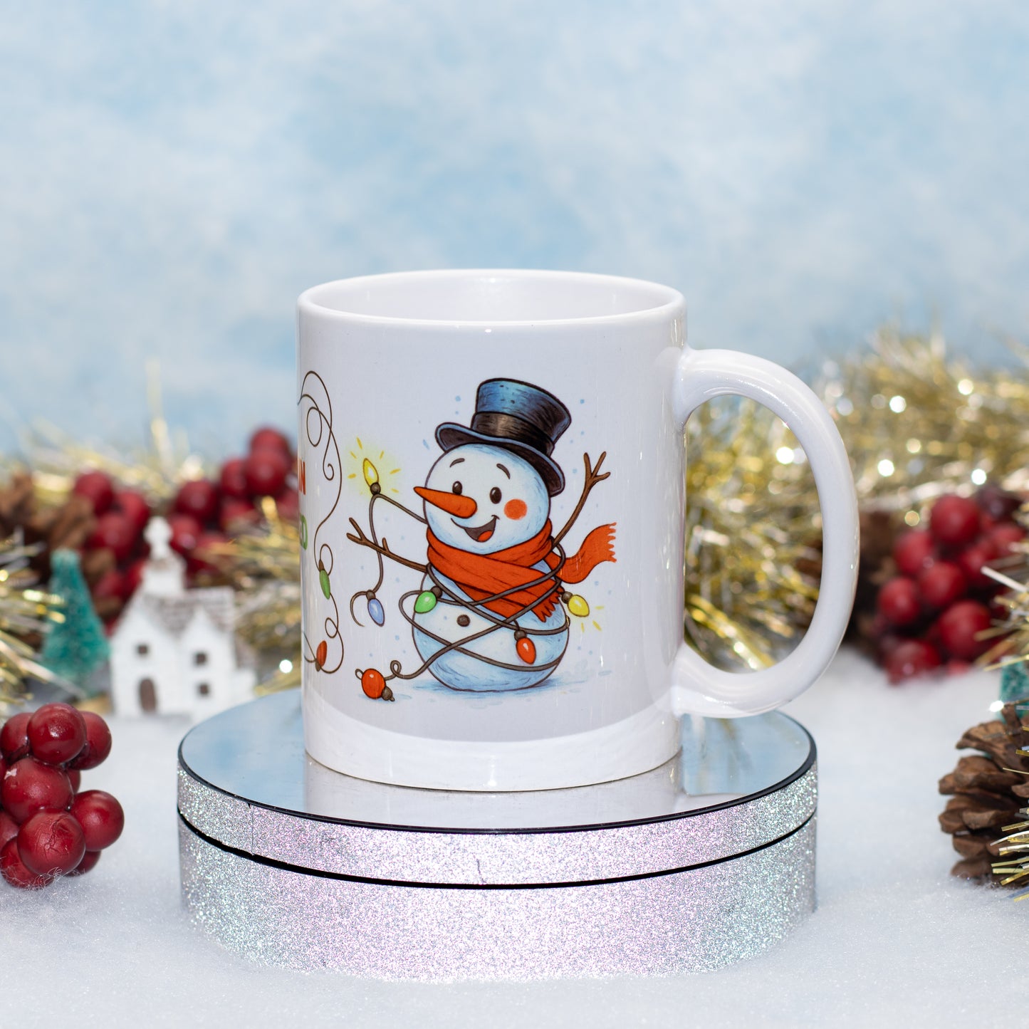 A white ceramic mug featuring a cartoon snowman illustration, displayed on a round glittery pedestal with festive holiday decorations in the background.
