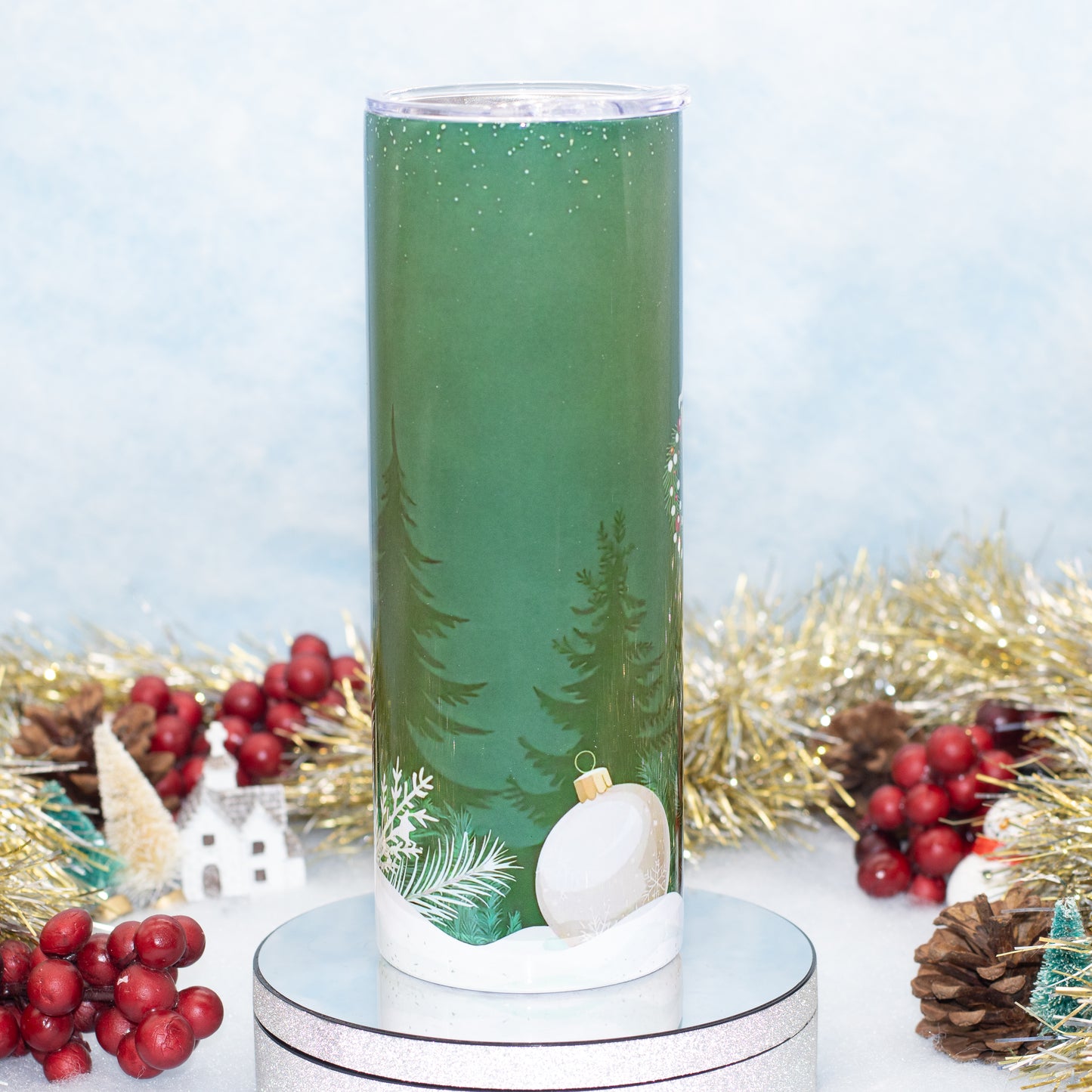 A tall green tumbler with a winter forest design, featuring pine trees, a white ornament, and snow, displayed on a silver base with festive holiday decorations in the background.