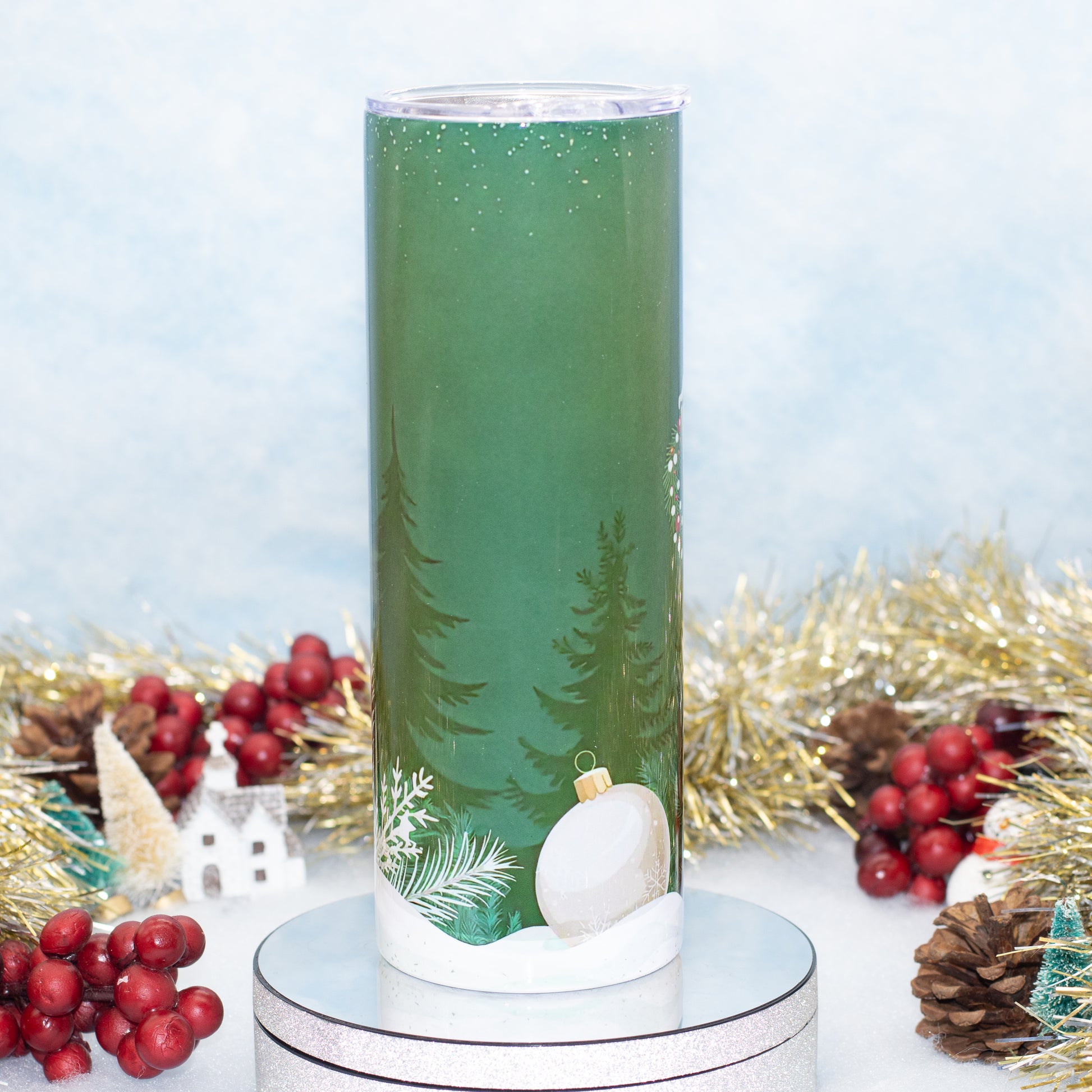 A tall green tumbler with a winter forest design, featuring pine trees, a white ornament, and snow, displayed on a silver base with festive holiday decorations in the background.