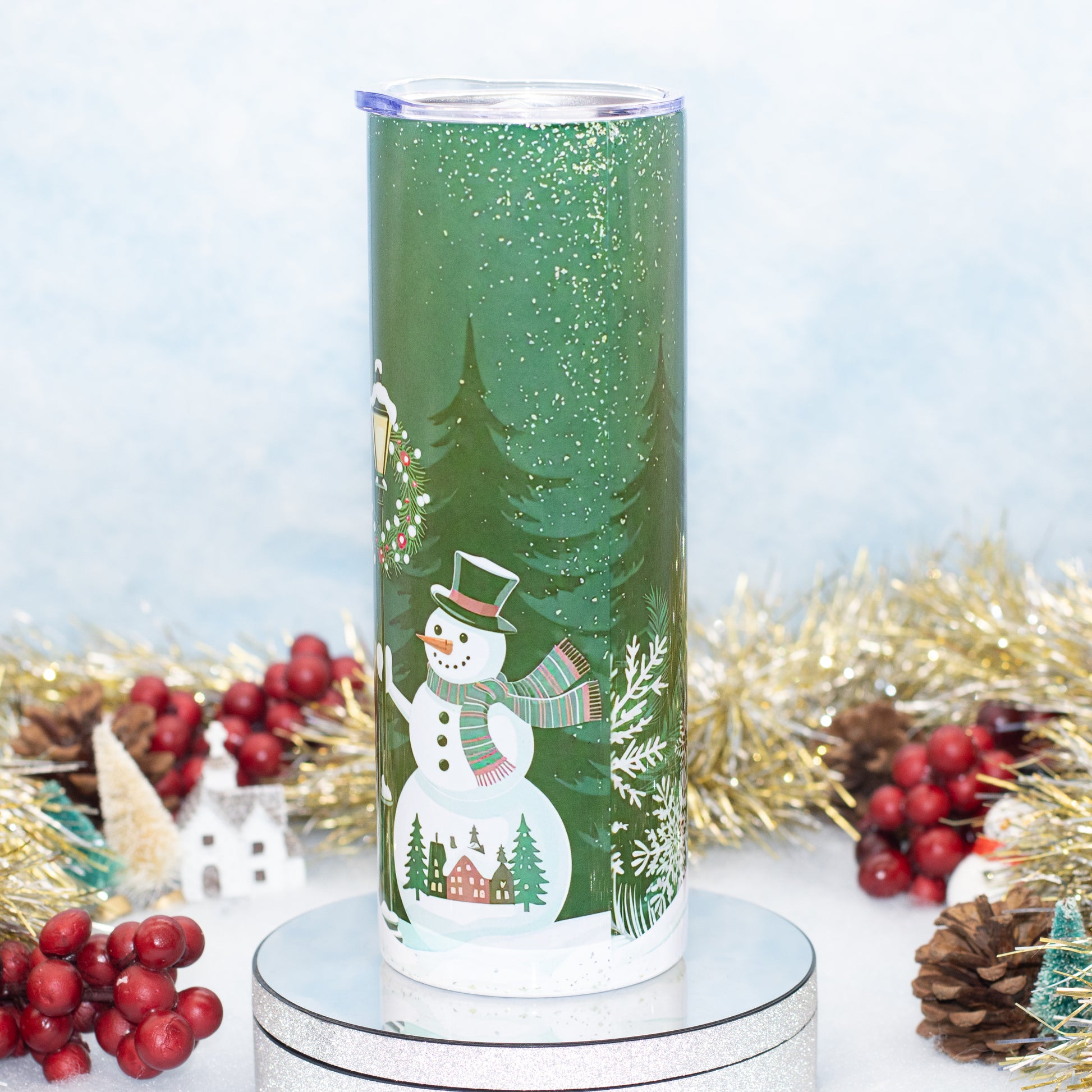 A tall insulated tumbler with a festive winter holiday design, featuring a snowman, evergreen trees, and a small house on a snowy ground. The tumbler is decorated with green glitter and has a clear lid, shown standing on a silver display base with holiday decorations like tinsel, pinecones, and red berries in the background.