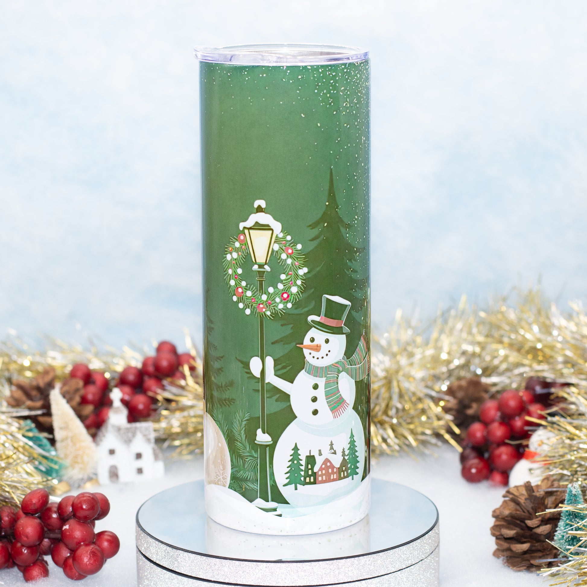 A tall green tumbler with a winter holiday scene printed on it, featuring a snowman, snow lamp post, evergreen trees, and small houses. The tumbler is shown on a round silver pedestal with festive tinsel, pinecones, and red berries around it against a soft light blue background.
