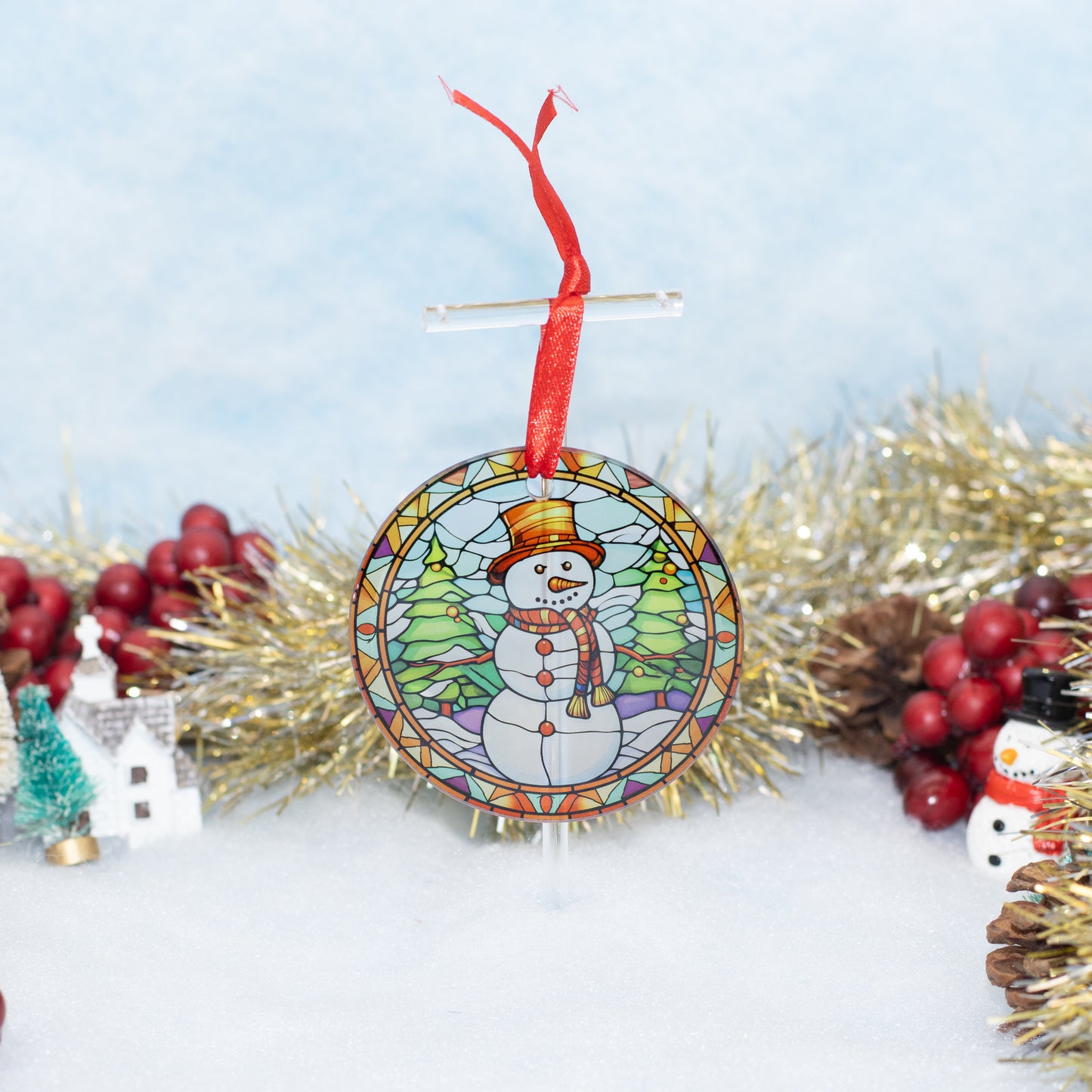A round stained-glass-style Christmas ornament featuring a snowman wearing a top hat and scarf among green trees, hanging from a red ribbon. The ornament is displayed on a white surface with festive tinsel, red berries, and small holiday decorations around it.