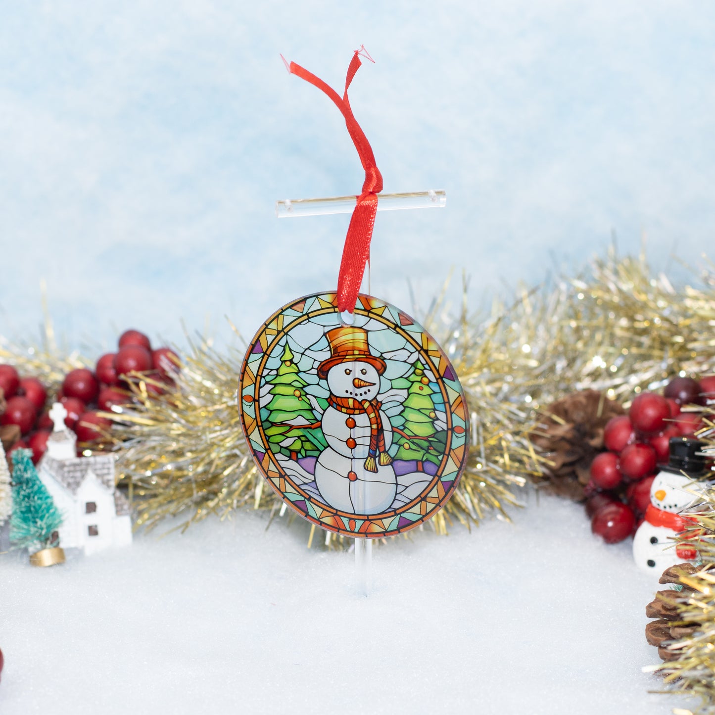 A round Christmas ornament featuring a stained-glass-style snowman illustration with a red hanging ribbon, displayed on a stand against a snowy white background with gold tinsel and holiday decorations.
