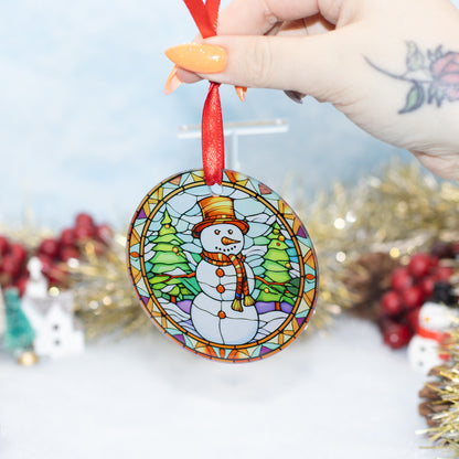 A hand holds a round stained-glass-style Christmas ornament featuring a snowman in a winter scene with trees. The ornament has a red hanging ribbon, and the background is softly blurred with holiday decorations.