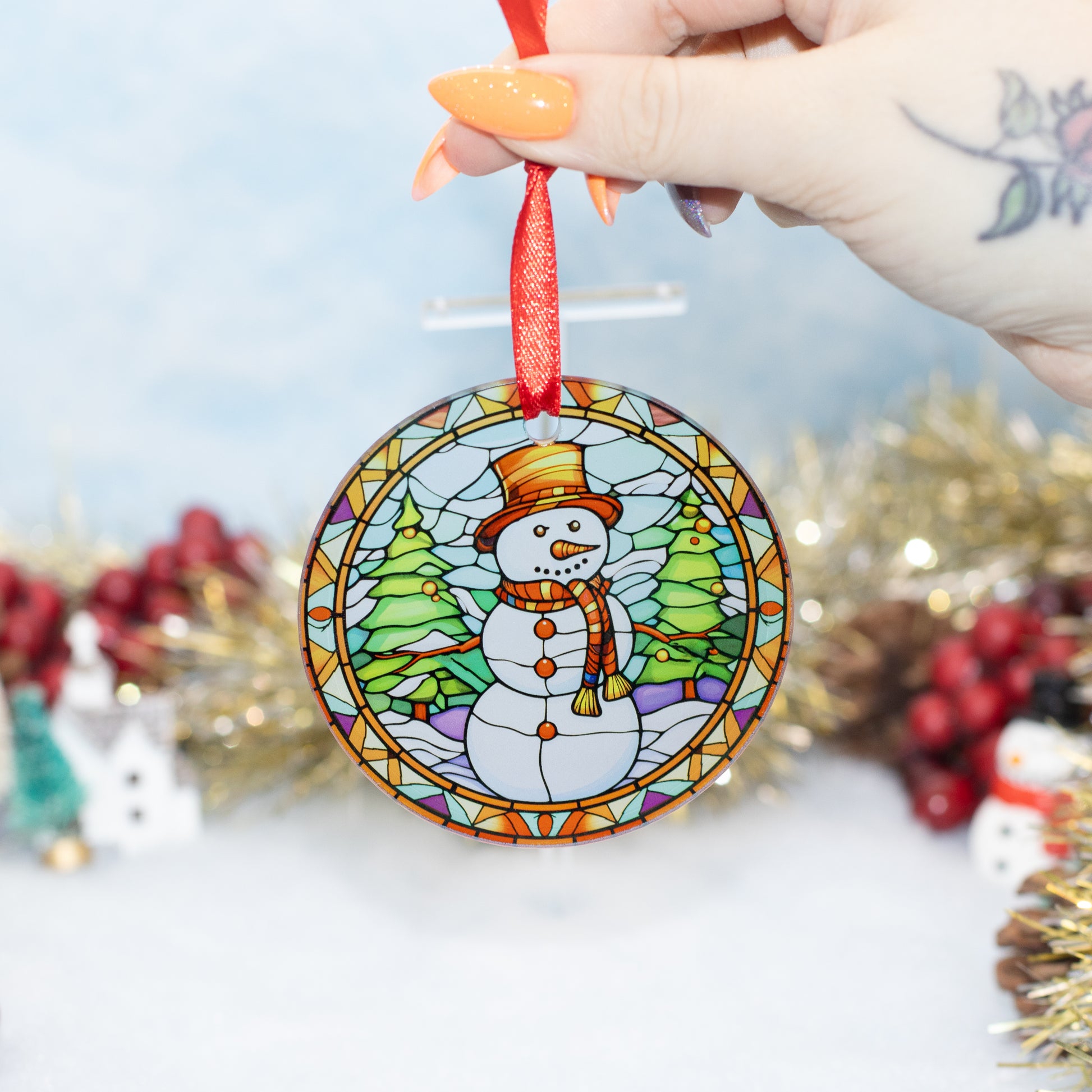 A hand holds a round stained-glass-style Christmas ornament featuring a snowman wearing a hat and scarf, with green trees and a red hanging ribbon. The background is softly blurred with holiday decorations.