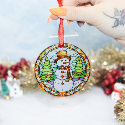 A hand holds a round stained-glass-style Christmas ornament featuring a snowman wearing a hat and scarf, with green trees and a red hanging ribbon. The background is softly blurred with holiday decorations.