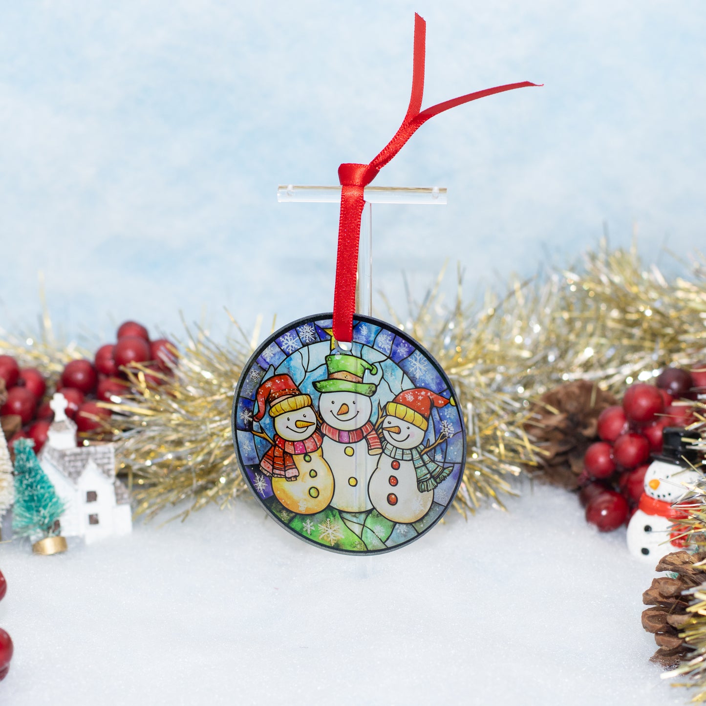 A round Christmas ornament featuring three snowmen in a stained-glass style, hanging from a red ribbon on a clear stand. The ornament is displayed on a white surface with festive holiday decorations, including gold tinsel, red berries, and miniature holiday props, against a soft light blue background.