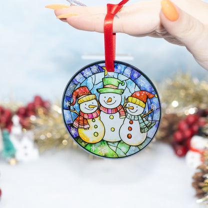 A round stained-glass-style Christmas ornament featuring three snowmen, held by a hand with orange nail polish against a softly blurred festive background.