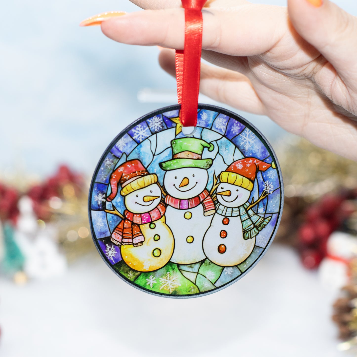 A round stained-glass-style Christmas ornament featuring three snowmen in colorful winter clothing, held by a hand with a red ribbon. The background is softly blurred with festive holiday decorations.
