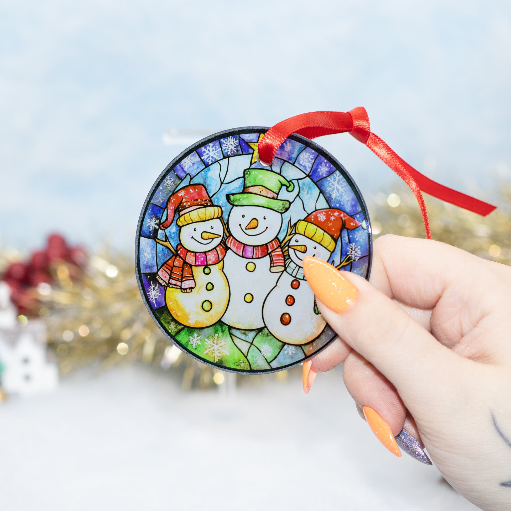 A hand holds a round stained-glass-style Christmas ornament featuring three snowmen wearing colourful hats. The ornament has a red ribbon hanging from the top and is photographed against a softly blurred festive background with gold tinsel and a light blue backdrop.