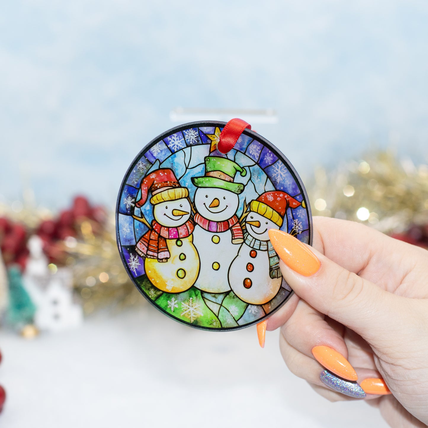 A hand holds a round stained-glass-style Christmas ornament featuring three snowmen in colourful hats and scarves, with a red hanging loop at the top. The background is softly blurred with holiday lights and decorations.