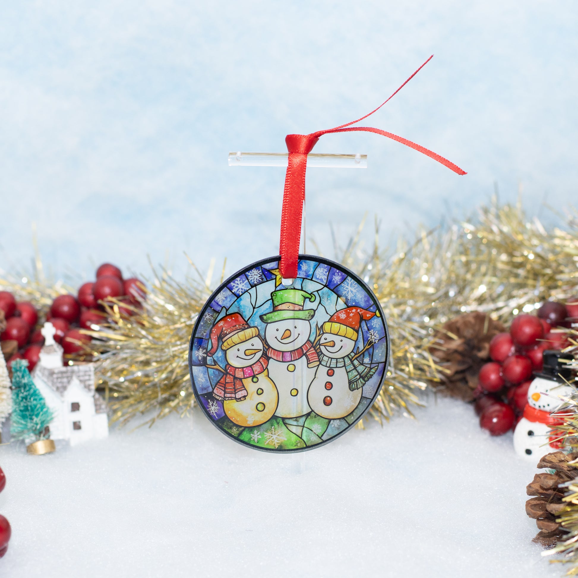 A round Christmas ornament featuring three snowmen in a stained-glass style design, hanging from a red ribbon on a clear stand. The ornament is displayed on a white surface surrounded by gold tinsel, red berries, and small holiday decorations against a soft light blue background.