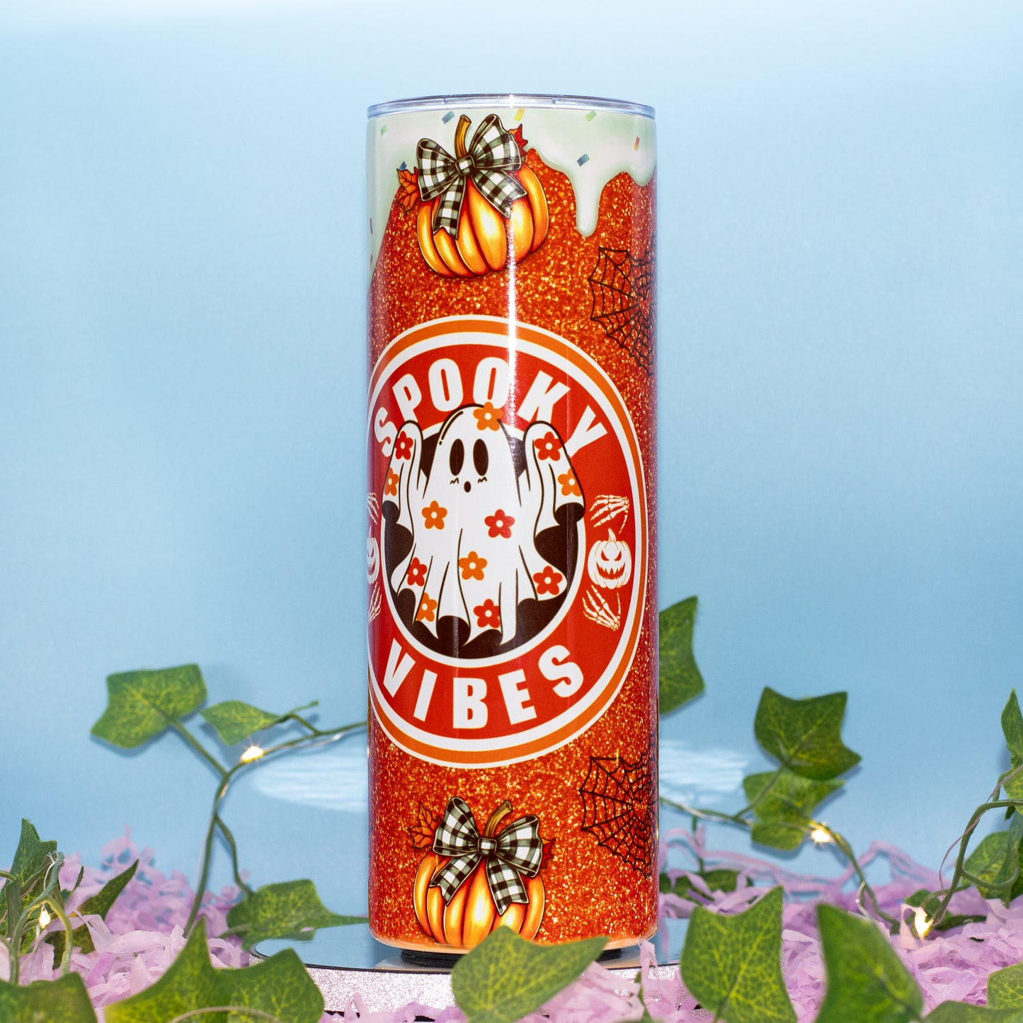 A tall cylindrical tumbler with a glittery orange body, decorated with a white ghost, pumpkins, spiderwebs, and the words 'Spooky Vibes' in a red circular graphic. The tumbler is shown on a light blue background with decorative green ivy and purple flowers at the base.