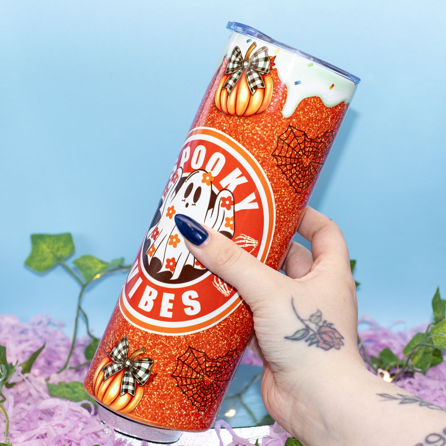 A hand holds a tall, slim tumbler decorated with a Halloween theme. The tumbler features orange glitter, pumpkins with plaid bows, spiderwebs, a ghost, and the words 'Spooky Vibes' in a circular logo.