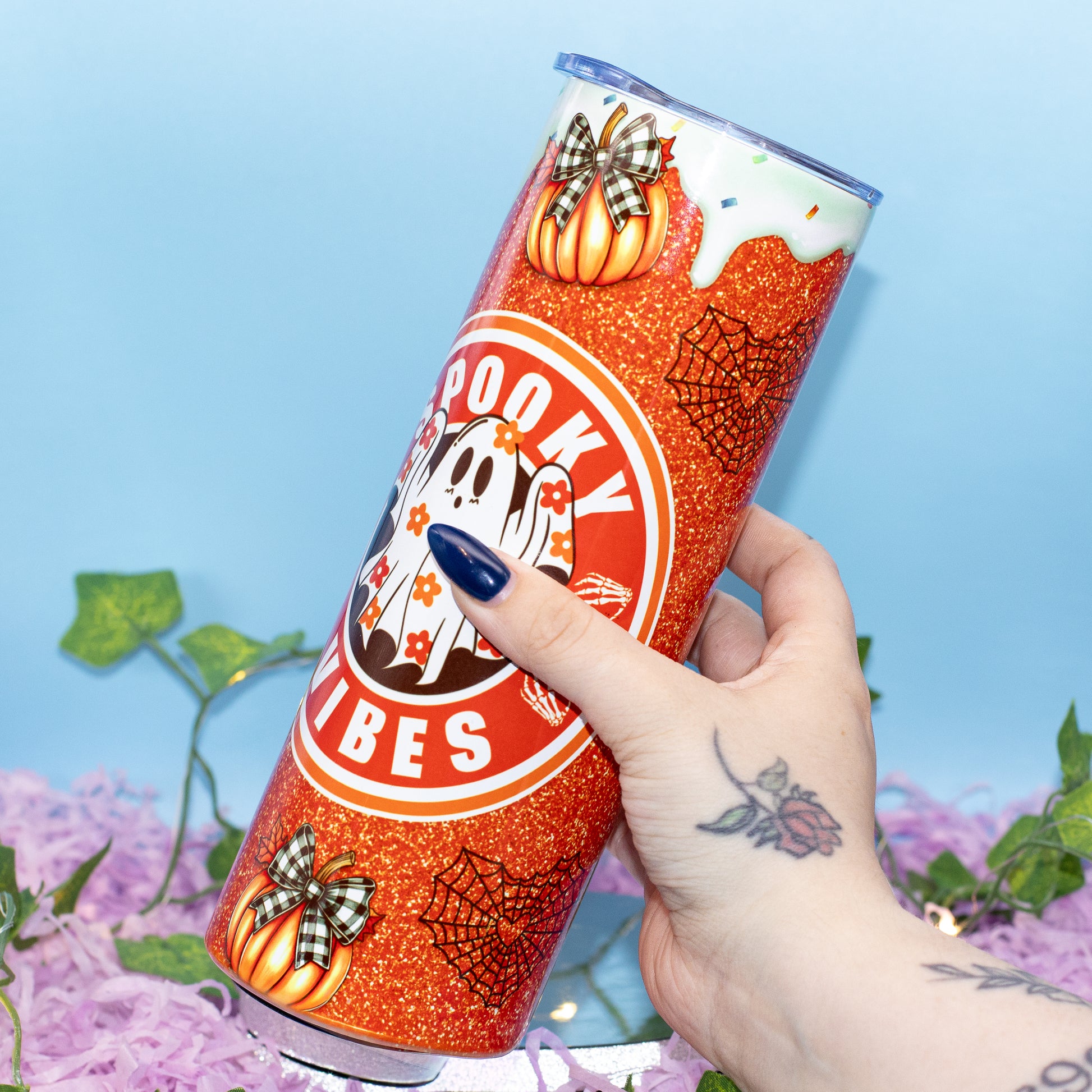 A hand holds a tall, slim tumbler decorated with a Halloween theme. The tumbler features orange glitter, pumpkins with plaid bows, spiderwebs, a ghost, and the words 'Spooky Vibes' in a circular logo.