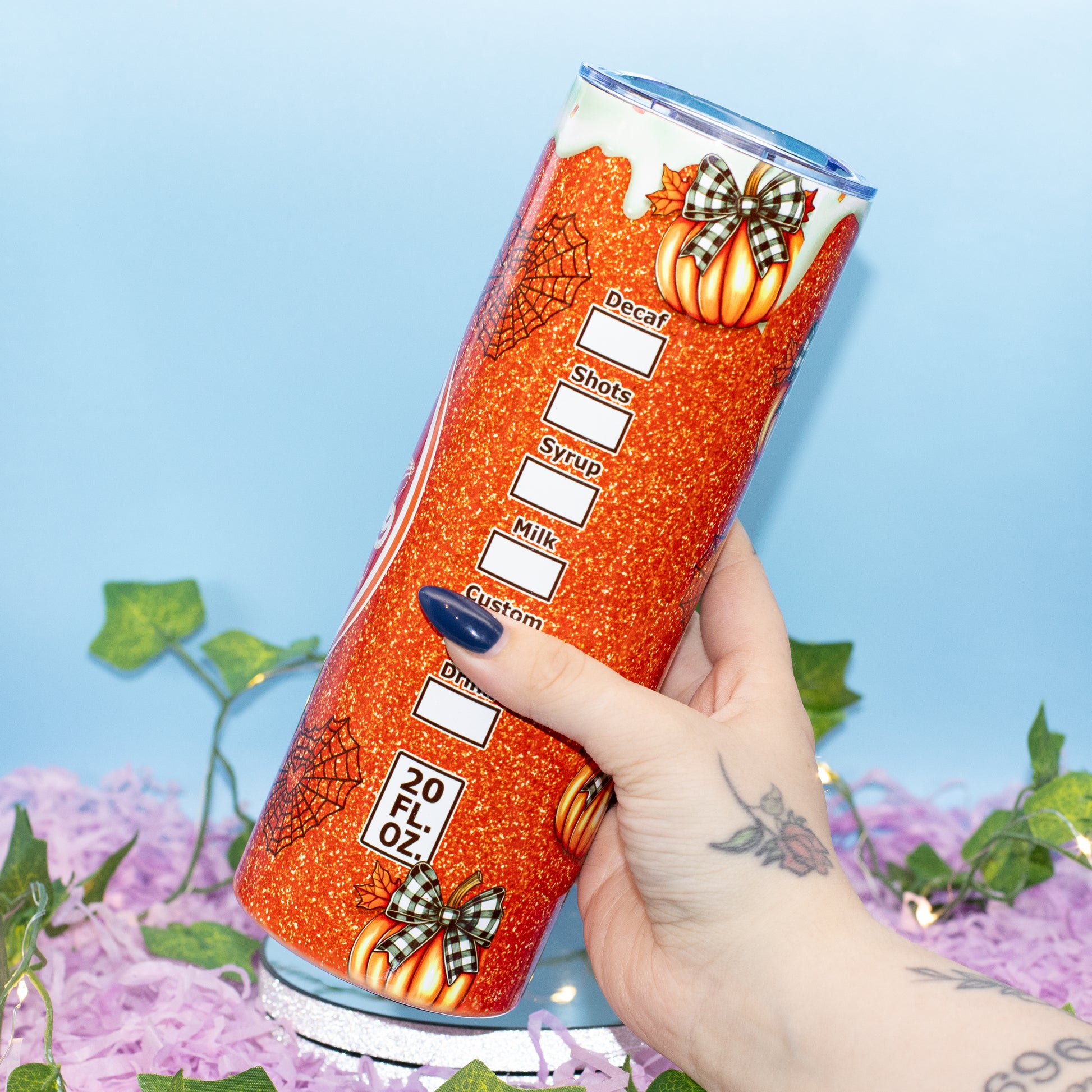 A hand holds a tall, cylindrical tumbler decorated with orange glitter, pumpkins, spiderwebs, and a pumpkin patch graphic. The tumbler features white text fields for ingredients like Decaf, Shots, Syrup, Milk, and Cream.