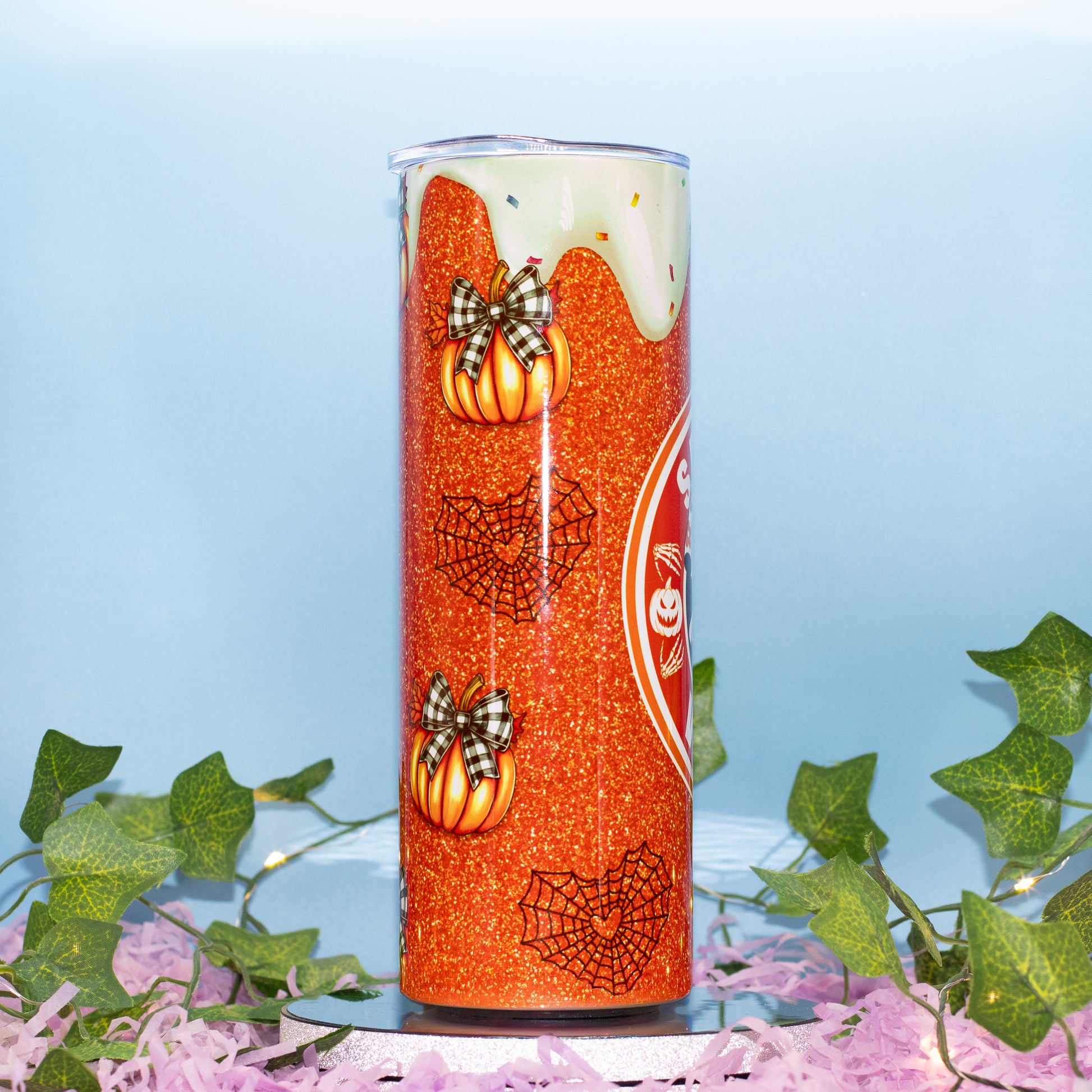 A tall tumbler with a Halloween-themed orange glitter wrap, featuring orange pumpkins with plaid bows, black spiderwebs, and a white dripping design near the top. The tumbler is displayed on a silver base surrounded by pink shredded paper and green ivy against a light blue background.