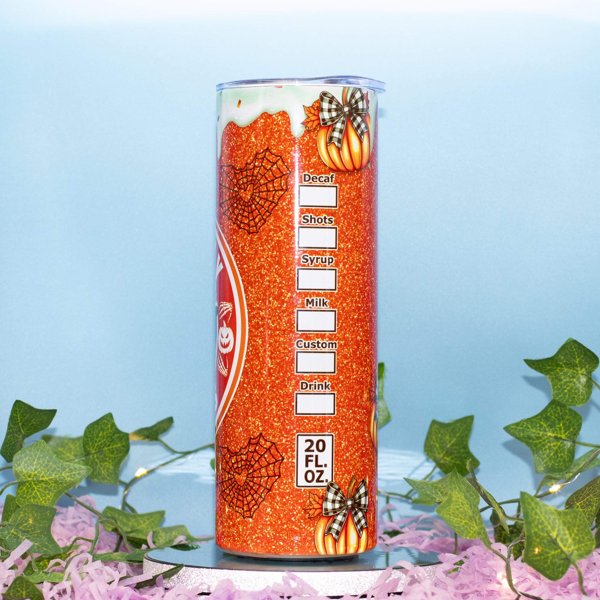 An orange glitter tumbler with Halloween-themed graphics, including spiderwebs, pumpkins, and a pumpkin patch, shown on a light blue background with decorative green ivy and pink shredded paper at the bottom.